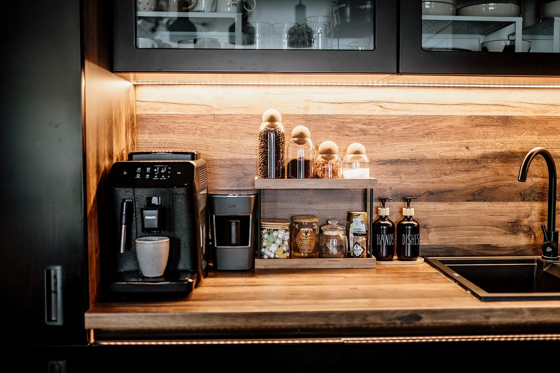 Cabinet and counter feature a coffee station with machines for brewing and grinding, jars of coffee, and decorative bottles. The setting shows a tidy kitchen space. Planning a coffee station starts with the sequence, not the shelf