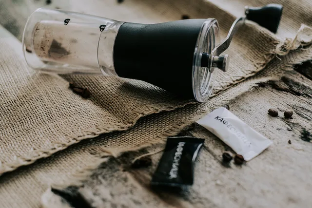 A coffee grinder sits on a burlap cloth alongside small packets of coffee. Coffee beans are scattered nearby, creating a cozy coffee-making scene that highlights brewing essentials. 5 parts of home brewing costs, explained