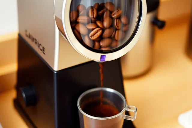 Burr coffee grinder grinding beans into a dosing cup on a counter. How to find coffee gear worth owning as a beginner: the useful version
